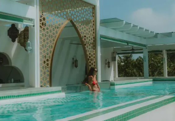 Woman in red bikini relaxing in a turquoise rooftop pool by a decorative arched screen, white pergola, and tropical greenery under a sunny sky