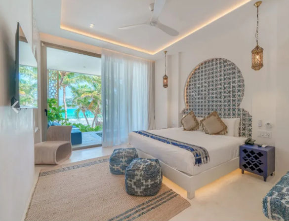 Sunlit tropical beachfront resort bedroom with king bed, Moroccan-style tiled headboard, hanging lanterns, blue poufs and sliding glass doors opening to palm trees and turquoise ocean view.