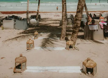 Tropical beach with antique-style lanterns lining a white-painted sandy path between palm trees toward turquoise sea and lounge cabanas