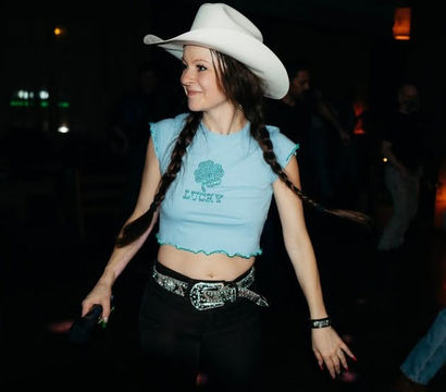 Person in a white cowboy hat with braided pigtails dancing in a dimly lit country bar, wearing a light blue cropped 'Lucky' top and sparkly belt — lively indoor nightlife scene.