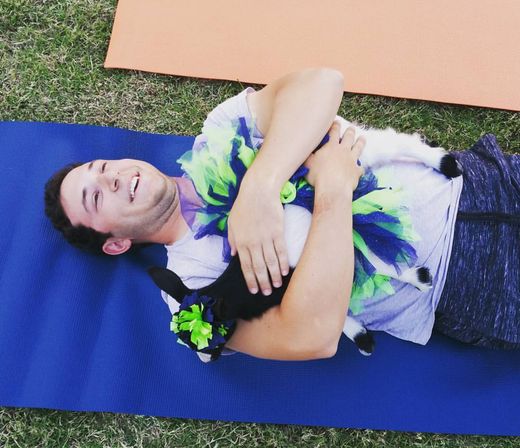 Smiling man lying on a blue yoga mat on grass, hugging a black-and-white baby goat wearing a green-and-blue tutu and flower headpiece, playful outdoor goat yoga moment