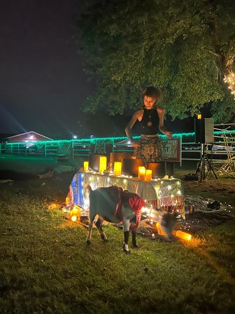 Nighttime outdoor wellness event in a grassy field — a performer plays crystal singing bowls at a candlelit, string-lit table while a goat in a red bandana grazes nearby.