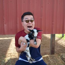 Person in sunglasses holding a black-and-white baby goat in front of a red metal barn, seated on hay at a sunny petting-zoo farm.