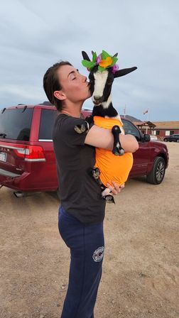 Person kissing a small black-and-white goat wearing an orange shirt and a colorful flower crown, held in a dusty rural parking lot beside a red SUV and flags under a cloudy sky