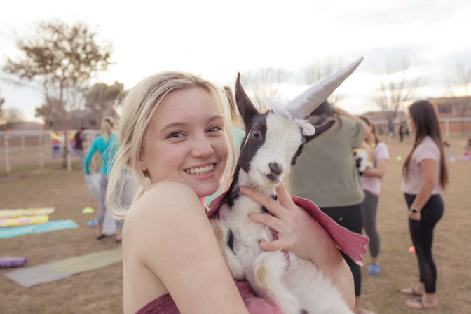 Smiling young woman holding a black-and-white baby goat dressed as a unicorn with a horn and pink cape at an outdoor goat yoga event in a park