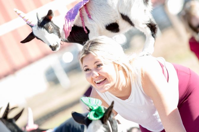 Smiling woman in an outdoor goat yoga class with a small goat wearing a unicorn horn and purple wings balancing on her back