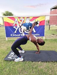 Woman in outdoor goat yoga backbend on a mat holding a small goat in a denim vest, grassy field with colorful banner and fence