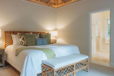 Spa-like coastal bedroom with rattan headboard and bench, crisp white bedding, green accent pillows, warm bedside lamps, and an open doorway to a bright ensuite bathroom with a glimpse of tropical greenery.