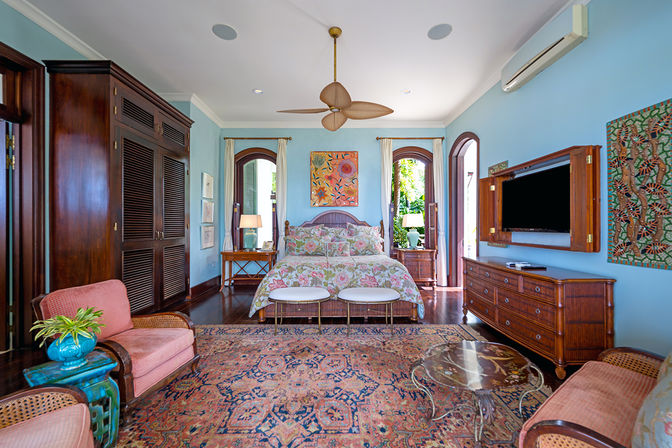 Sunlit tropical bedroom with light blue walls, floral-patterned bedding on a rattan bed, wooden dresser and wardrobe, arched windows opening to garden greenery, woven ceiling fan, pink upholstered chairs and ornate area rug.