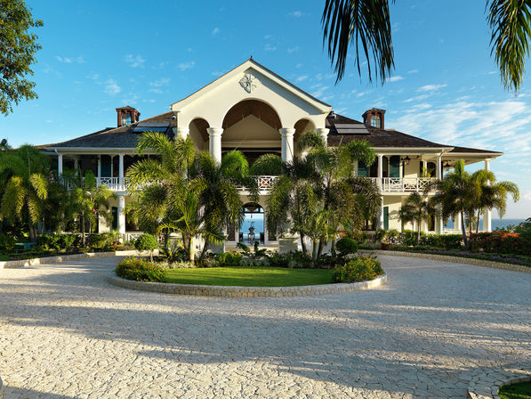 Oceanfront tropical villa with white columns and arched central entry, flanked by palm trees and a circular stone driveway under a bright blue sky.