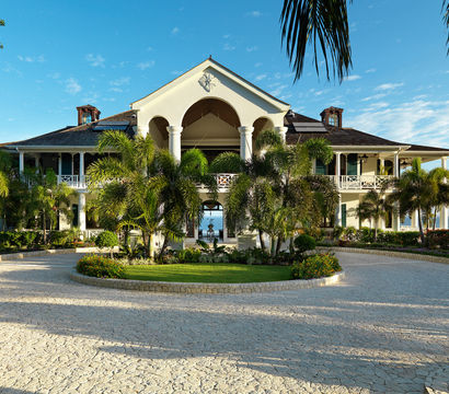 Oceanfront tropical villa with white columns and arched central entry, flanked by palm trees and a circular stone driveway under a bright blue sky.