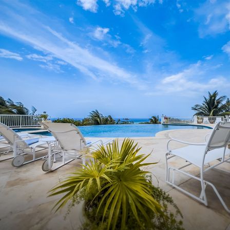 Oceanfront infinity pool overlooking turquoise sea with white lounge chairs, tropical palms and a bright blue sky with wispy clouds