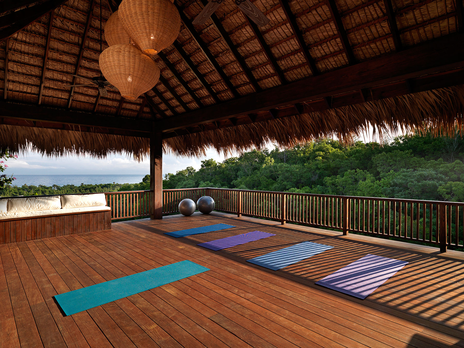 Tropical open-air yoga pavilion on a wooden deck with colorful mats, exercise balls, wicker pendant lights, and ocean-and-forest view.