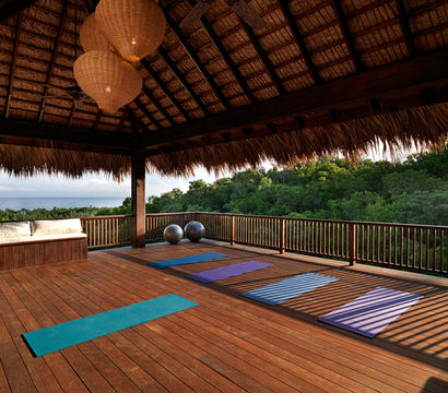 Tropical open-air yoga pavilion on a wooden deck with colorful mats, exercise balls, wicker pendant lights, and ocean-and-forest view.