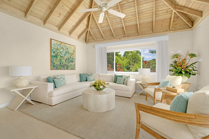 Bright, breezy coastal-style living room with vaulted exposed wood ceiling and ceiling fan, white L-shaped sectional with aqua pillows, round textured coffee table, rattan armchairs, indoor plants and a large window framing lush tropical greenery.