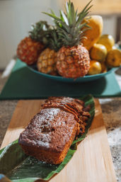 Sliced banana bread loaf dusted with powdered sugar on a banana leaf and wooden board, with a blurred bowl of pineapples and mangoes for a tropical kitchen vibe.