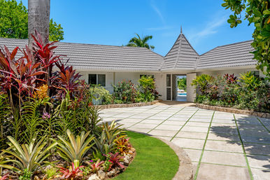 Sunny tropical villa courtyard with gray tiled roof and pointed turret, paved driveway with grass joints, lush landscaping of bromeliads, red ti plants and palms, and a glimpse of a pool under a bright blue sky