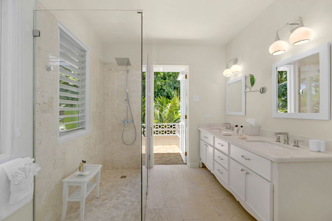 Sunlit coastal bathroom with glass-enclosed walk-in shower, white double-sink vanity, marble tile floor, and an open door leading to tropical palm foliage.