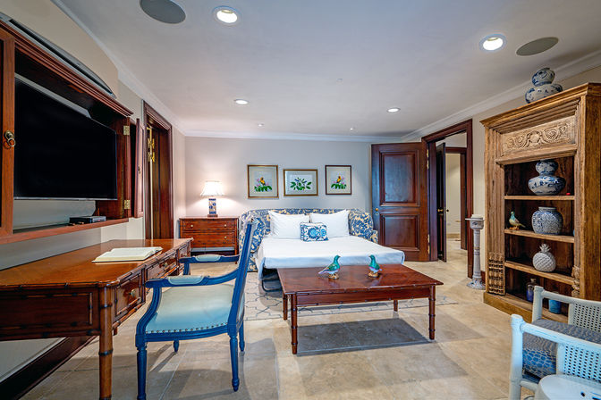 Bright coastal-inspired hotel suite living area with a white sofa bed, blue-accent armchairs, wooden desk and coffee table, carved wooden bookshelf with ceramic vases, tiled floor, and framed botanical prints.