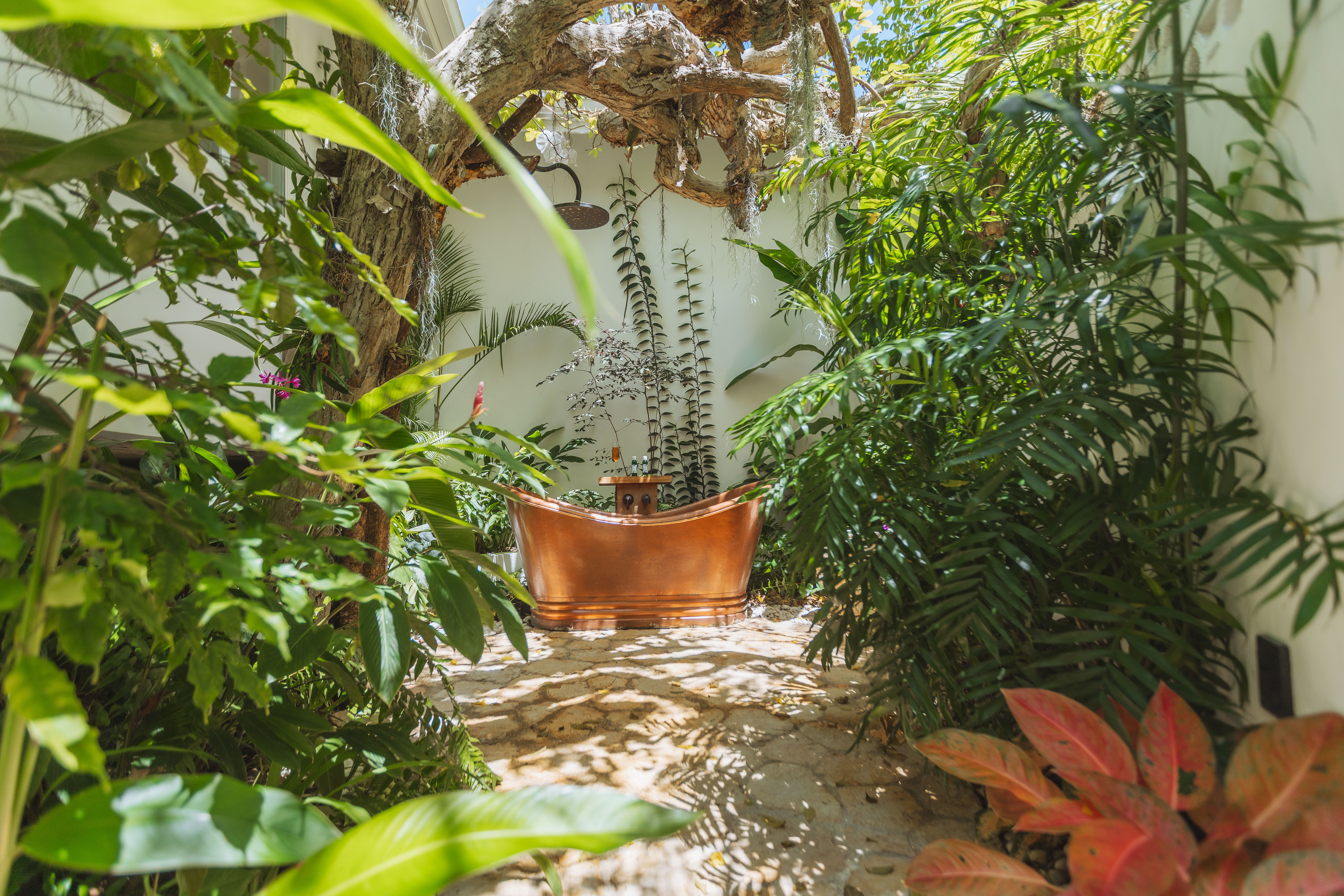 Sunlit tropical courtyard with a copper outdoor bathtub and showerhead, surrounded by lush green plants and a stone floor
