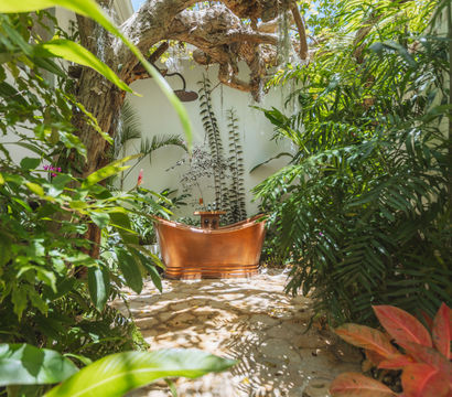 Sunlit tropical courtyard with a copper outdoor bathtub and showerhead, surrounded by lush green plants and a stone floor