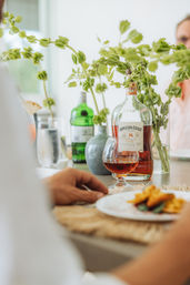 Cozy sunlit dining table close-up with a rum bottle and snifter of amber spirit, green vases holding leafy stems, a plate of appetizers, and a hand reaching toward the table.