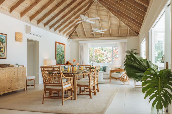 Airy open-plan coastal living and dining room with vaulted wood ceiling, rattan dining set, large windows and lush tropical plants