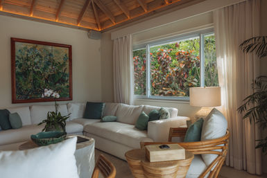 Sunlit tropical living room with a white sectional sofa, green accent pillows, rattan chairs, wooden vaulted ceiling and a large window framing lush garden foliage.