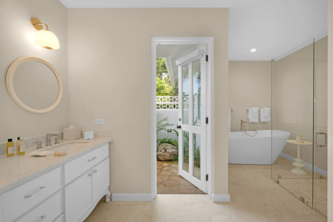 Bright neutral luxury bathroom with marble double vanity and round mirror, glass-enclosed shower, freestanding tub, and French doors opening to a small garden courtyard