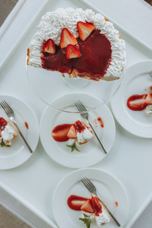 Top-down view of a sliced strawberry-topped cheesecake with whipped cream and glossy strawberry sauce, served on white plates on a white serving tray.