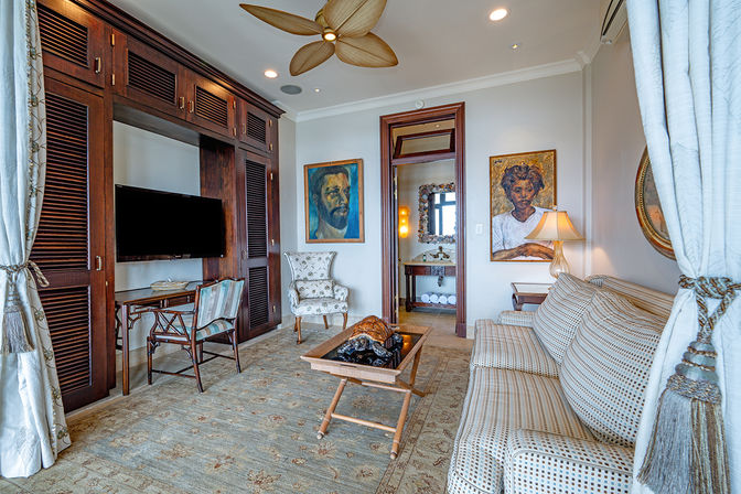 Bright coastal-style hotel suite living room with leaf-blade ceiling fan, dark wood built-in media wall and flat-screen TV, patterned sofa and floral armchair, wooden coffee table, framed portrait paintings and doorway to a bathroom with towels visible.