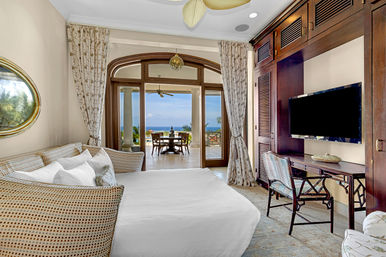 Tropical coastal bedroom suite with white bedding and wicker daybed, wooden built-in media desk and TV, and arched glass doors opening to a covered terrace dining area with ocean and palm views.