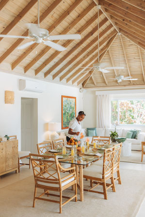 Bright tropical villa dining area with vaulted wood ceiling and white ceiling fans; rattan chairs around a glass table set for breakfast with orange juice and pastries, a host arranging the table beside a sunny living room and garden-view window.
