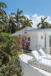 Sunny tropical villa patio with white cushioned lounge chairs and rolled turquoise towels on a balcony, surrounded by palm trees, red tropical plants and a bright blue sky.