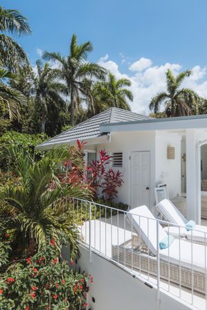 Sunny tropical villa patio with white cushioned lounge chairs and rolled turquoise towels on a balcony, surrounded by palm trees, red tropical plants and a bright blue sky.