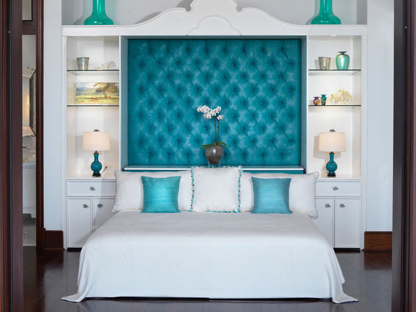 Bright luxury bedroom interior with large teal tufted headboard, crisp white bedding, teal accent pillows and lamps, built-in white shelving and a central orchid arrangement.