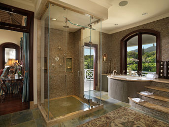 Luxurious spa-like bathroom with frameless glass rain shower, curved soaking tub on tiled steps, mosaic tile walls and arched window framing a lush hillside view.