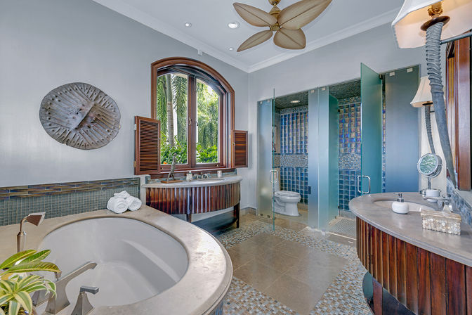 Resort-style tropical bathroom with a large soaking tub, curved wooden vanities, glass-enclosed toilet and shower with blue mosaic tile, arched window looking out to palm trees, and a leaf-shaped ceiling fan.