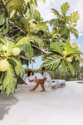 Two people relaxing on white wicker lounge chairs at a sunny tropical poolside terrace, shaded by a large tree with glossy lobed leaves and round green fruits for a lush outdoor vacation vibe.