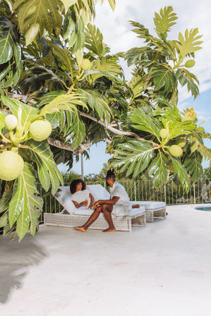 Two people relaxing on white wicker lounge chairs at a sunny tropical poolside terrace, shaded by a large tree with glossy lobed leaves and round green fruits for a lush outdoor vacation vibe.