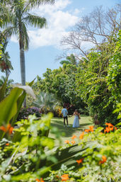 Couple holding hands strolling through a sunny tropical garden framed by tall palm trees, bright orange flowers and a clear blue sky.