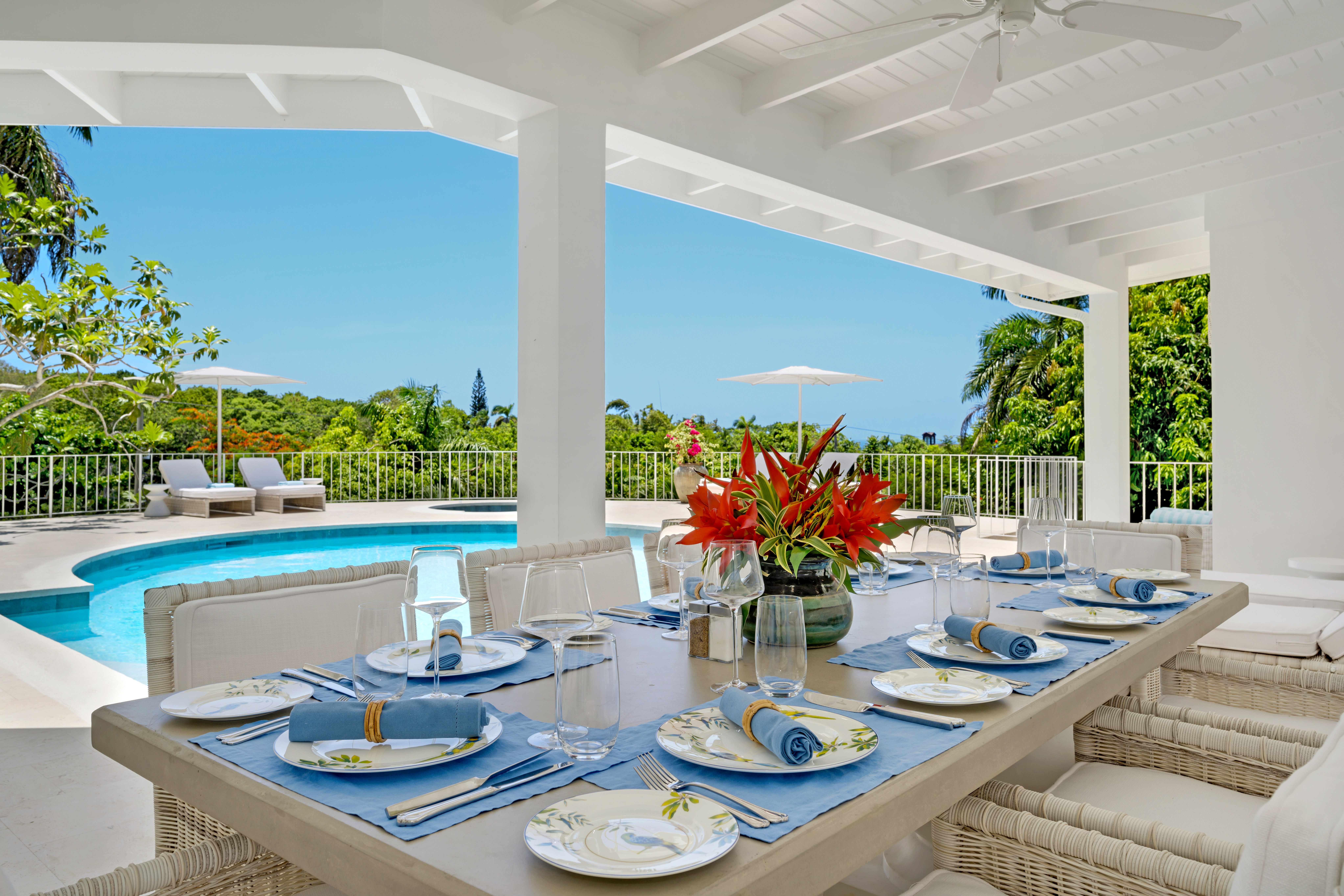 Sunny covered patio with a resort-style dining table set for eight—blue napkins, floral plates and a red tropical flower centerpiece—overlooking a curved swimming pool, loungers, white umbrella and lush palm-filled garden.