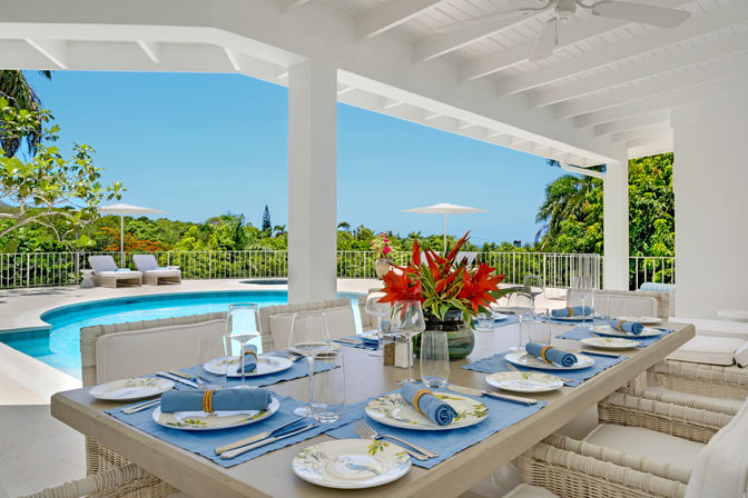 Sunny covered patio with a resort-style dining table set for eight—blue napkins, floral plates and a red tropical flower centerpiece—overlooking a curved swimming pool, loungers, white umbrella and lush palm-filled garden.
