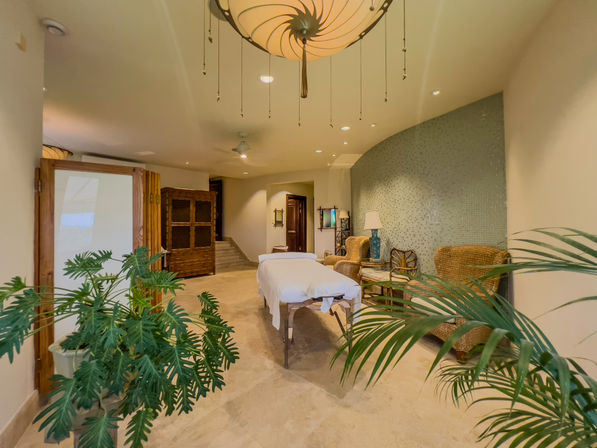 Resort-style spa treatment room with a central massage table draped in white linens, wicker lounge chairs, curved aqua mosaic wall, tropical potted plants, warm pendant lighting and neutral tile floor.