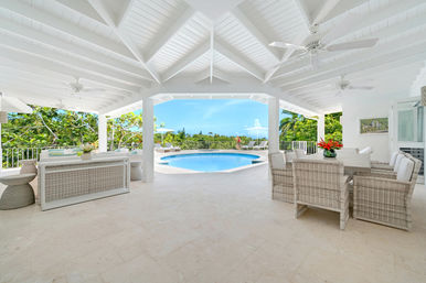 Sunny covered tropical villa patio with vaulted white ceiling and fans, wicker dining and lounge seating, tiled floor and round swimming pool overlooking lush greenery and blue ocean sky.