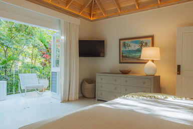 Sunlit tropical bedroom with vaulted wood ceiling, neutral decor, dresser and lamp, wall TV, and open doors to a balcony with a white wicker chair and lush garden—island getaway vibe.