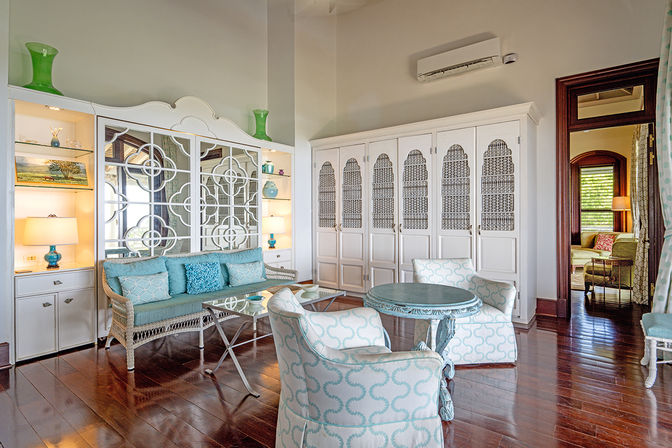Sunny coastal-style living room with turquoise accents: wicker sofa with blue cushions, patterned armchairs around a round painted table, mirrored decorative wall panels, built-in white shelving with lamps and vases, and polished dark hardwood floors.
