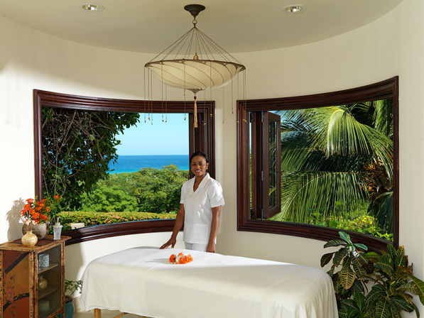 Ocean-view spa treatment room with a massage table, wide wooden-framed windows revealing tropical palms and sea, decorative plants and orange flowers, and a smiling spa therapist in a white uniform.