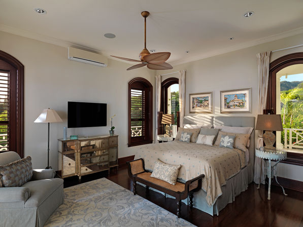 Sunlit tropical-style bedroom with king bed and patterned bedding, arched wooden shutters opening to lush greenery, wooden ceiling fan, mirrored dresser, wicker bench and cozy armchair.