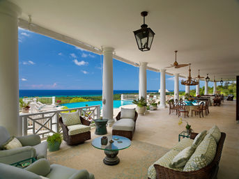 Oceanfront terrace with white columns, wicker lounge seating, infinity pool and panoramic turquoise sea view, plus al fresco dining under chandeliers and ceiling fans